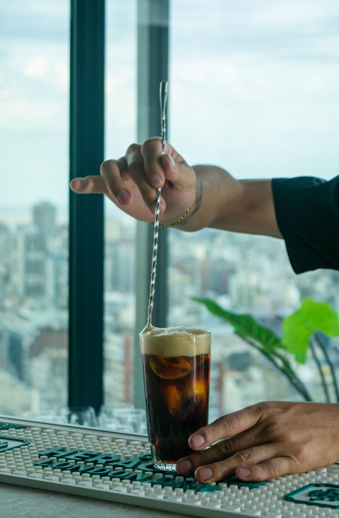 Barista mixing a cold brew coffee in a tall glass with a city skyline backdrop.