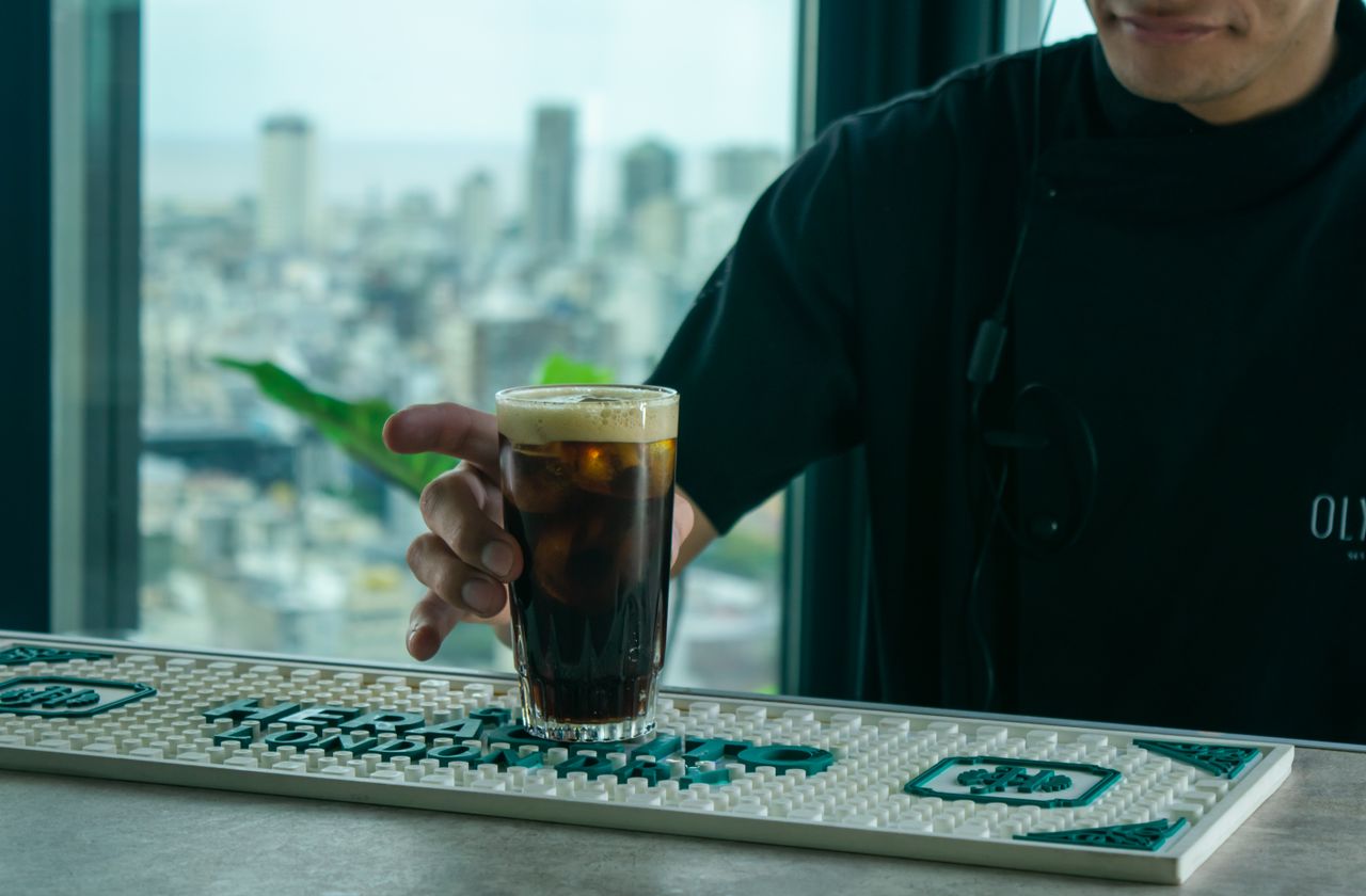 A barista serves a glass of iced coffee on a counter with a city skyline view.